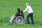 Betreuungskraft fährt eine pflegebedürftige Person im Rollstuhl